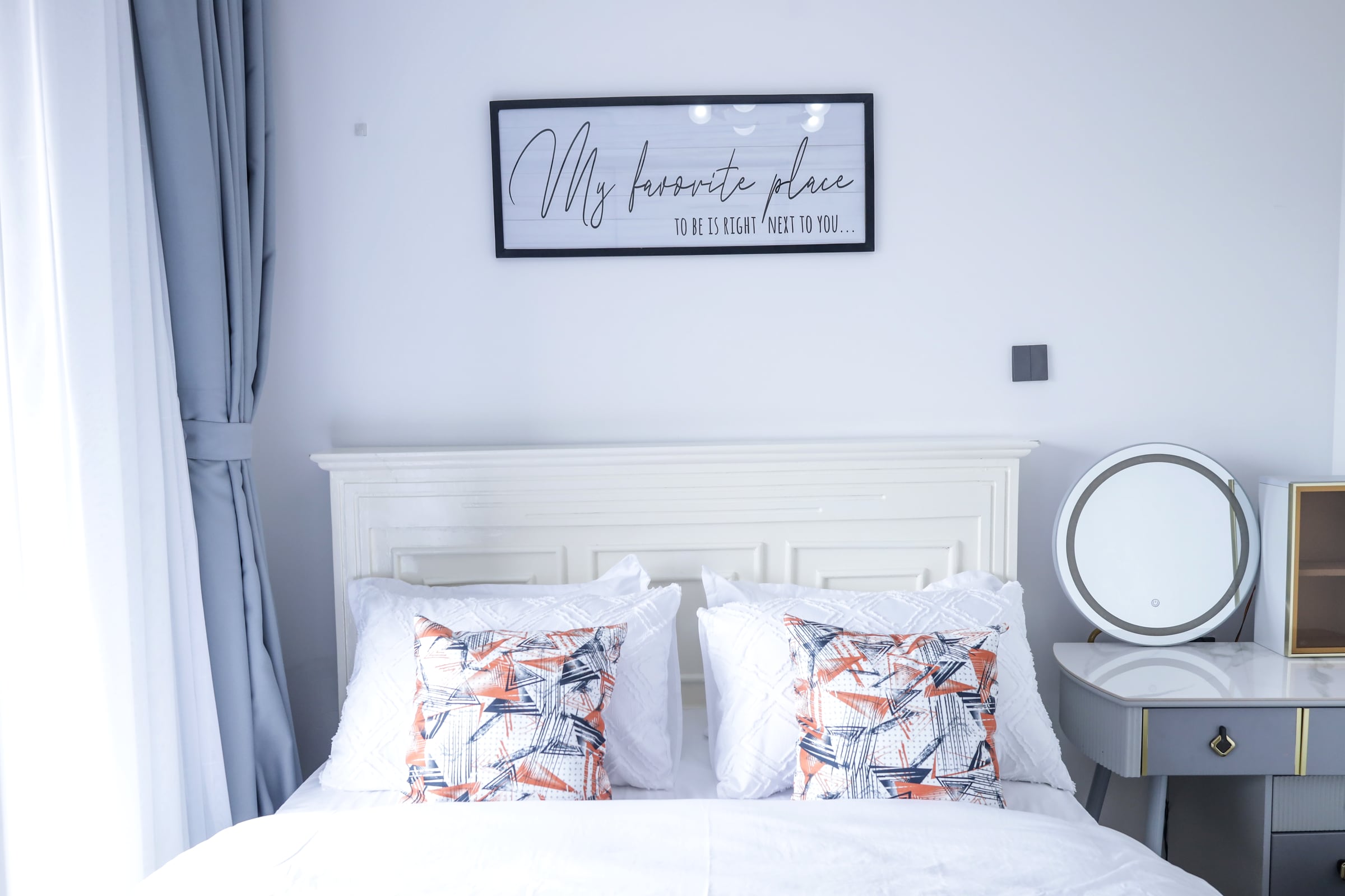 Bright bedroom with a white bed, round mirror and a globe chandelier under blown-out window light